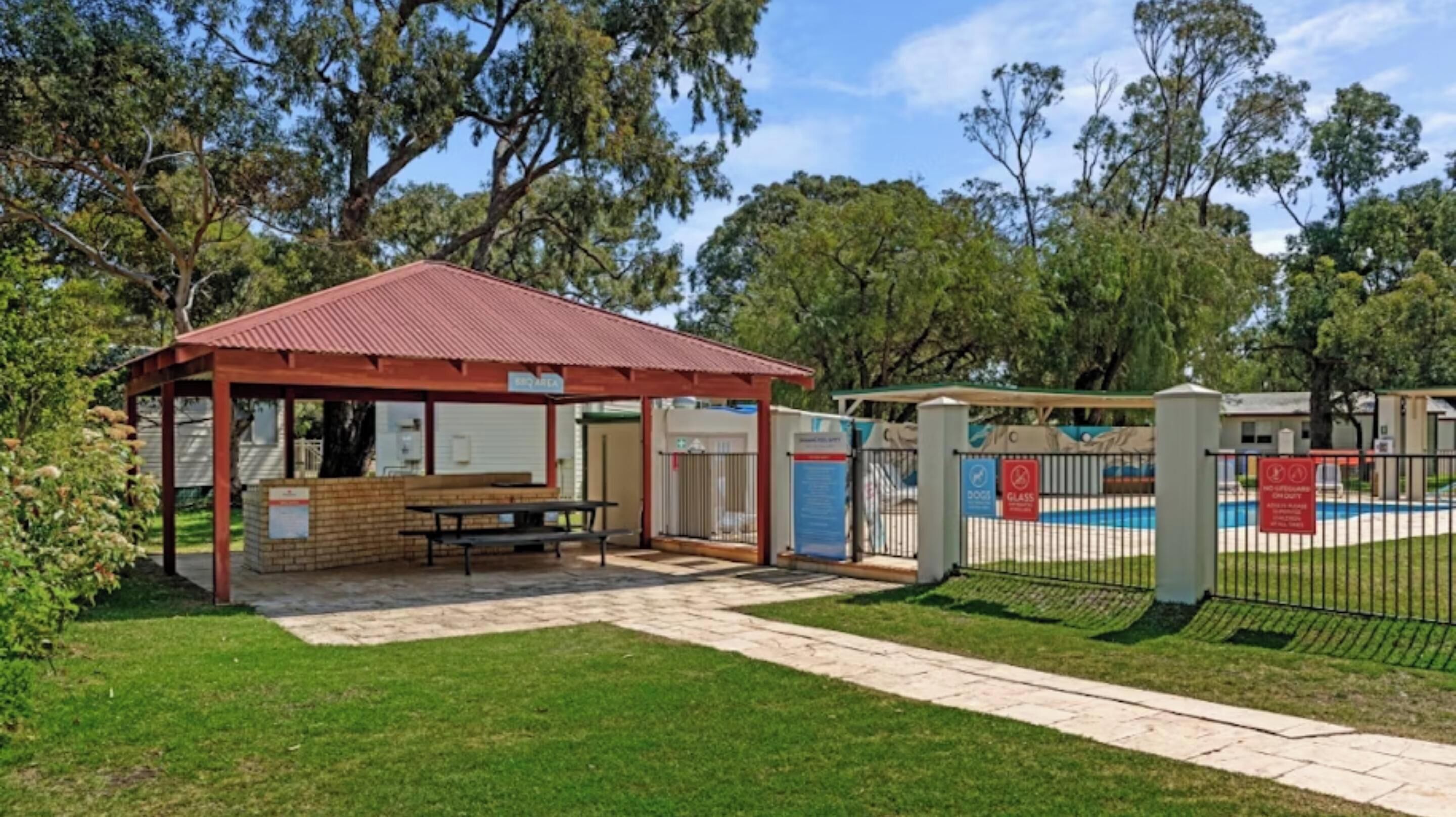 Un espace pique-nique abrité, équipé d’un banc et de tables de pique-nique, une piscine entourée d’une clôture et un bâtiment avec un toit rouge.