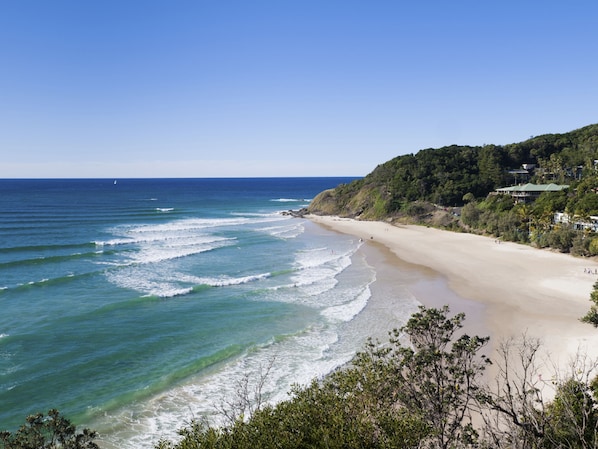 A beach with clear blue waters, a sandy shore, and a forested hillside in the background.