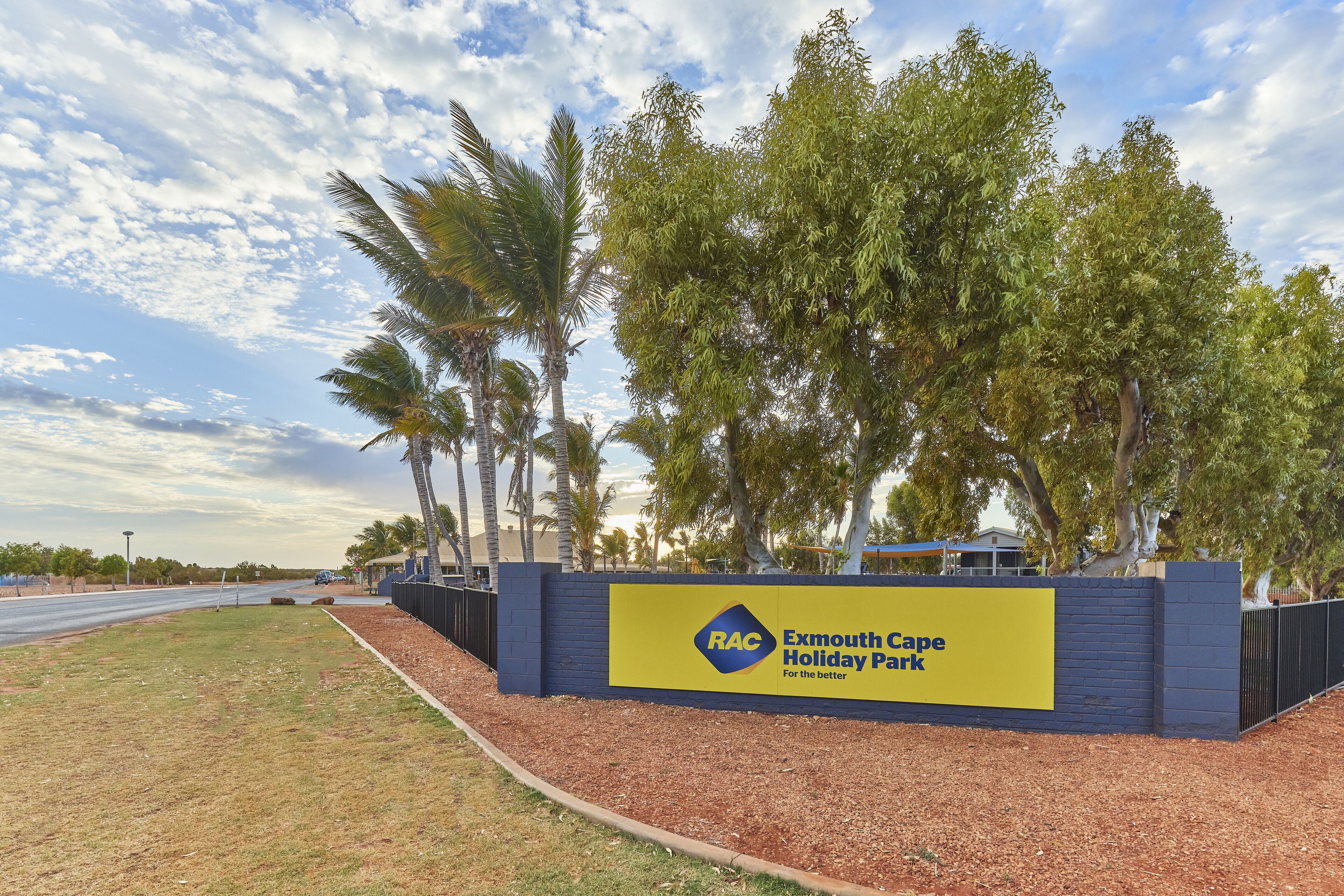 Señal para Exmouth Cape Holiday Park con un diseño de colores azul y amarillo, rodeada de palmeras y un cielo despejado.