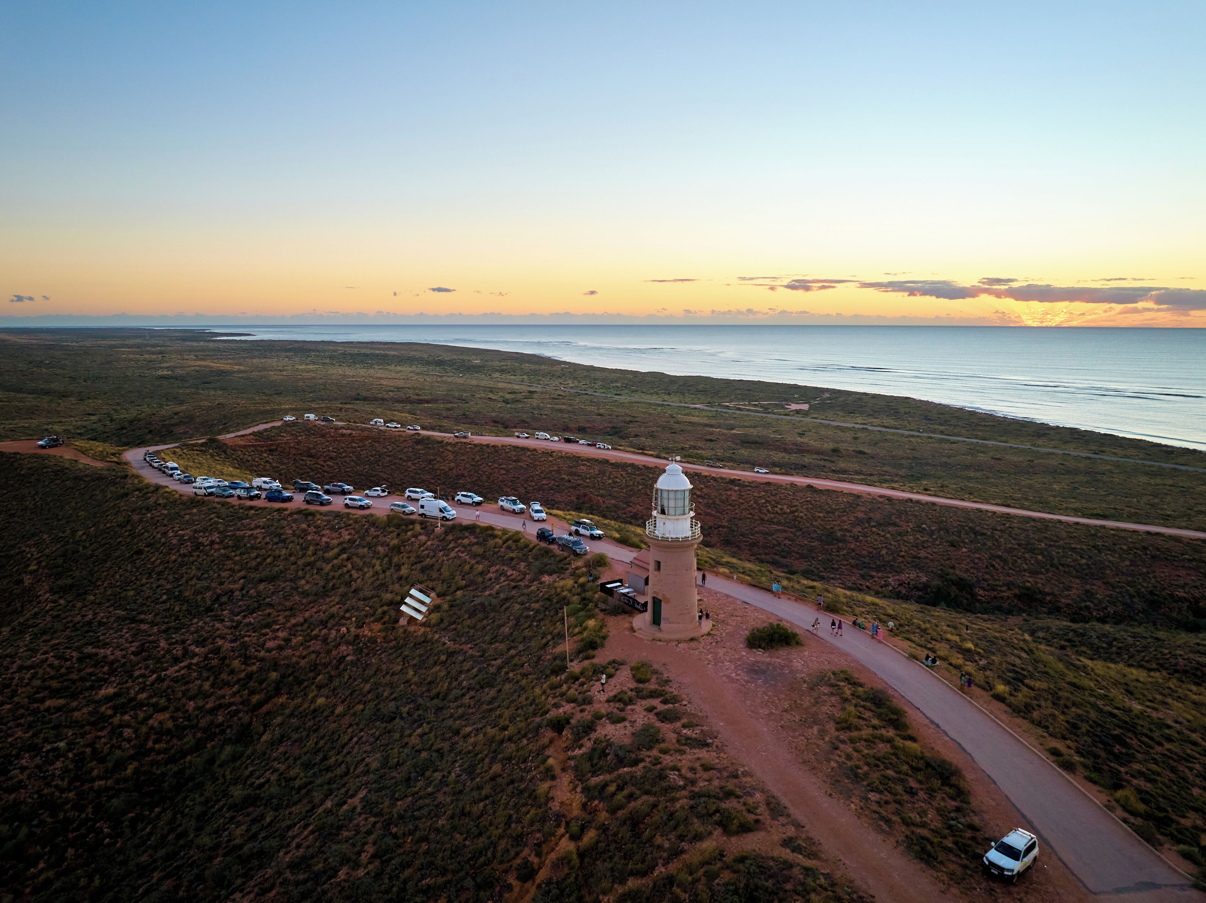 Vista aérea de una zona costera con un faro, un estacionamiento y una carretera que conduce a la playa.