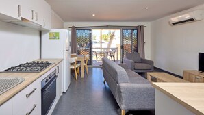 A modern kitchen with a gas stove, oven, and refrigerator, a dining area with a table and chairs, a gray sofa, and a TV on a wooden stand. - RAC Exmouth Cape Holiday Park (Exmouth)