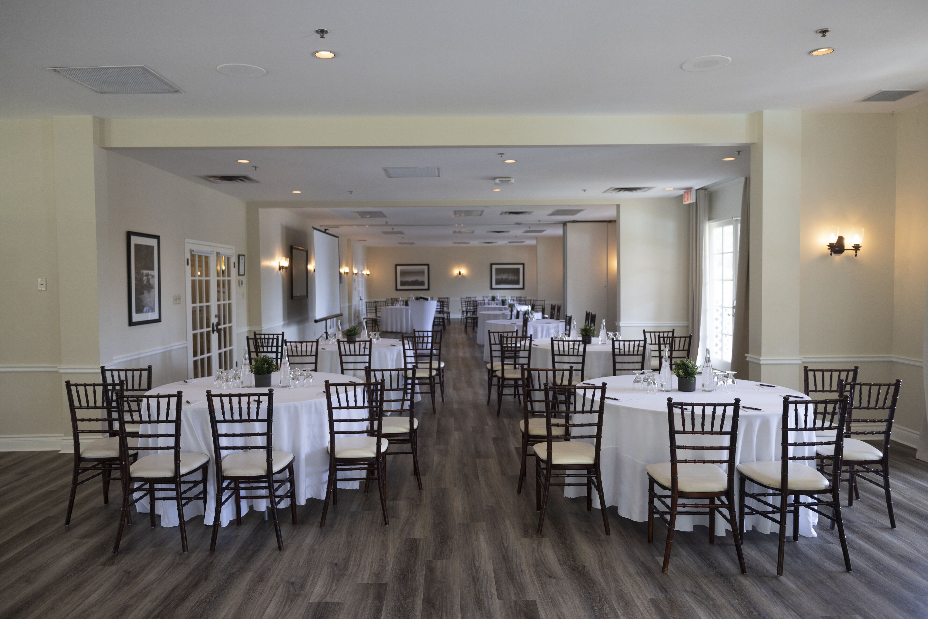 A long dining hall with multiple round tables and chairs, white tablecloths, and wall-mounted lighting.
