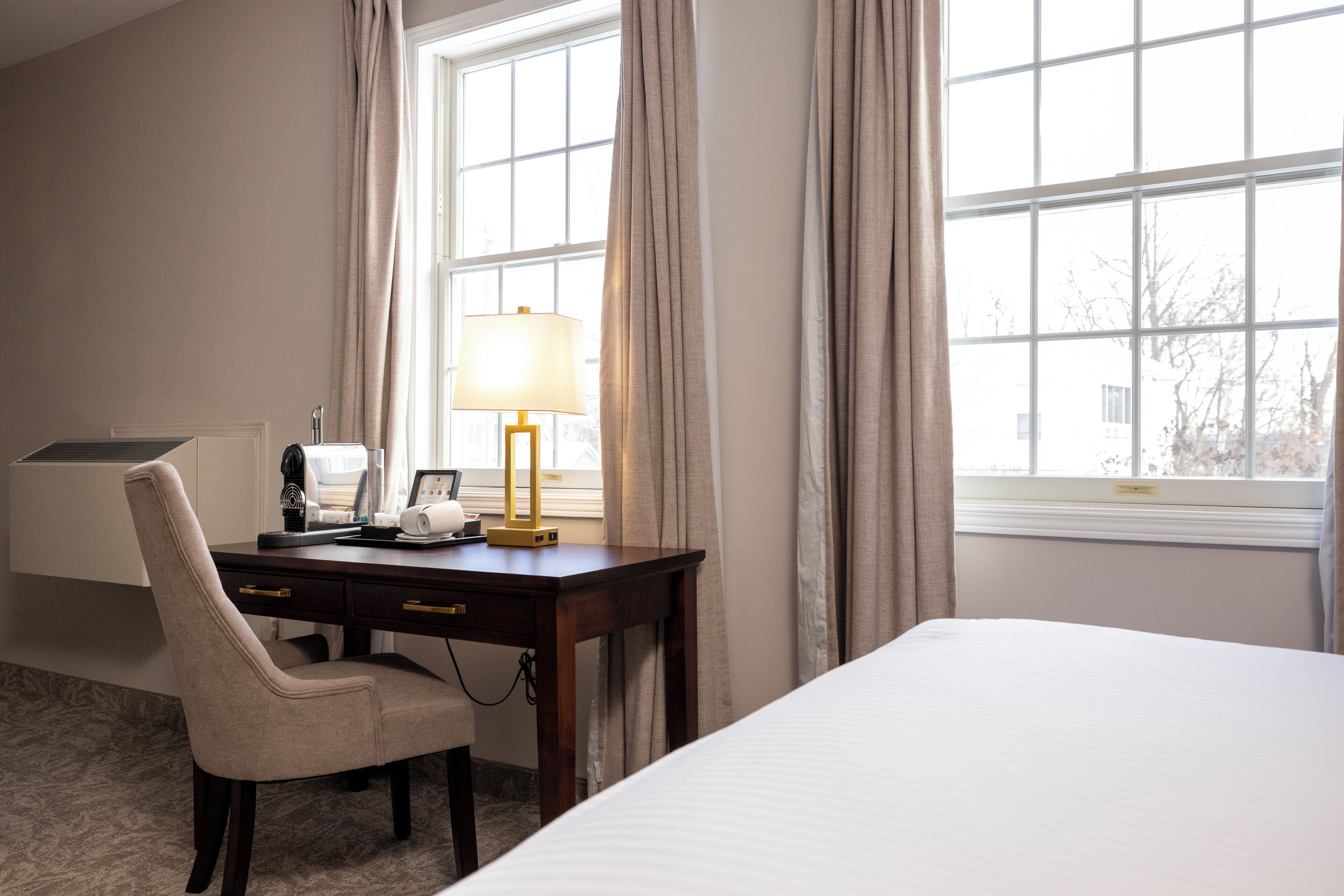 Una habitación de hotel con cama, escritorio con lámpara, silla y ventana con cortinas.