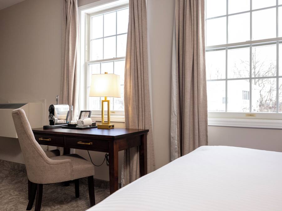 Una habitación de hotel con cama, escritorio con lámpara, silla y ventana con cortinas.