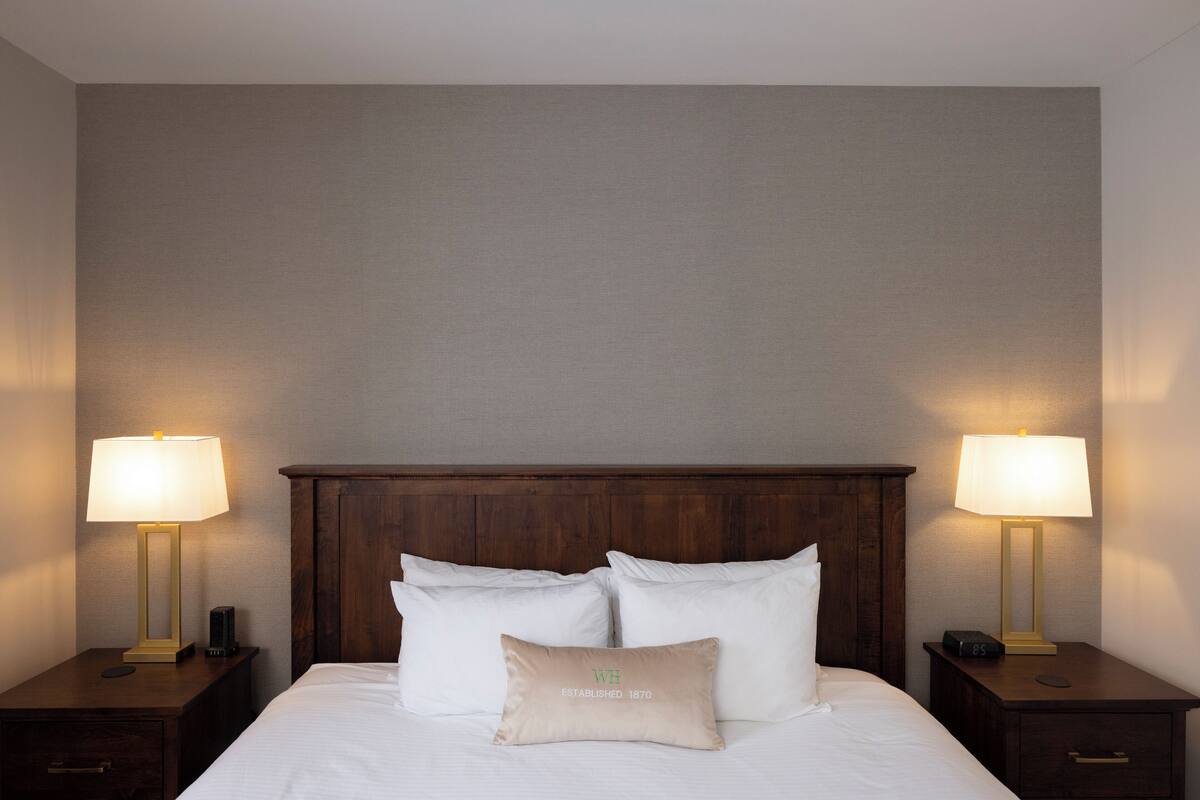 Un lit bien fait, avec du linge de lit blanc et un oreiller beige orné du texte « WH Established 1870 », flanqué de deux tables de chevet sur lesquelles sont posées des lampes et des réveils.