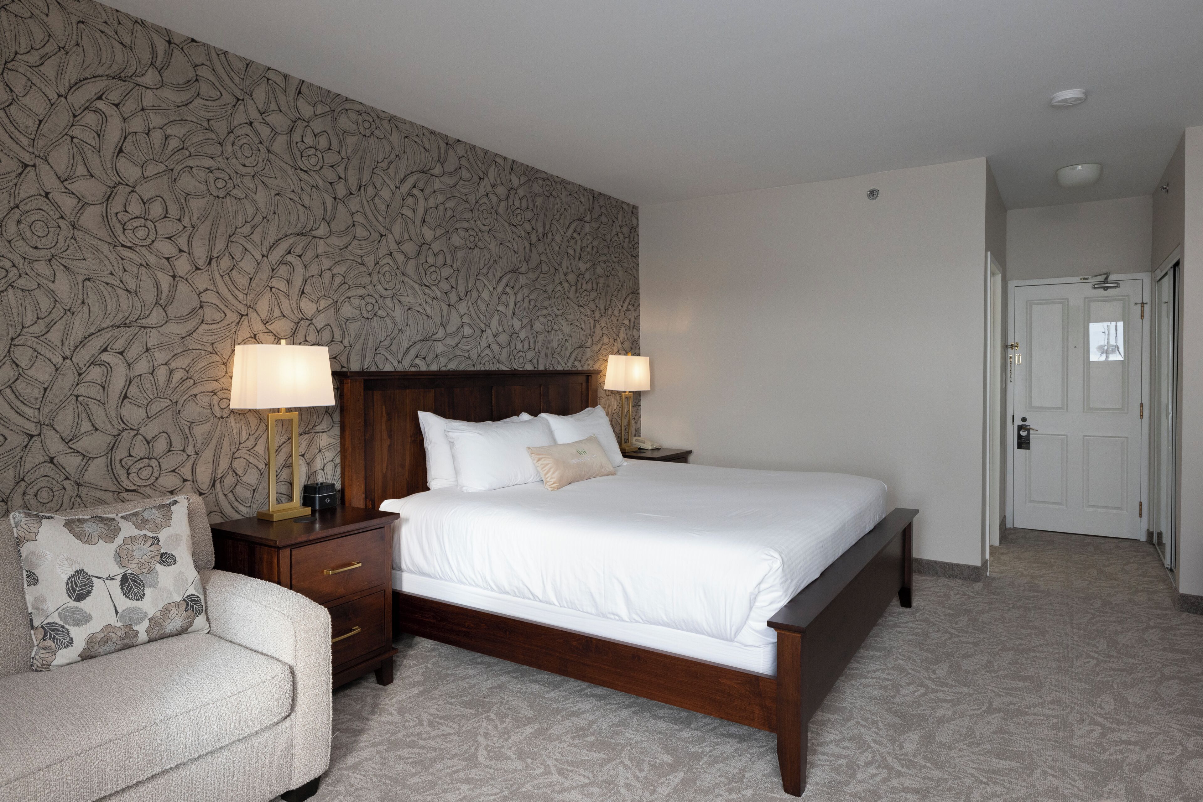 A neatly arranged hotel room with a large bed, two bedside tables with lamps, a wooden nightstand, and a floral-patterned wall.