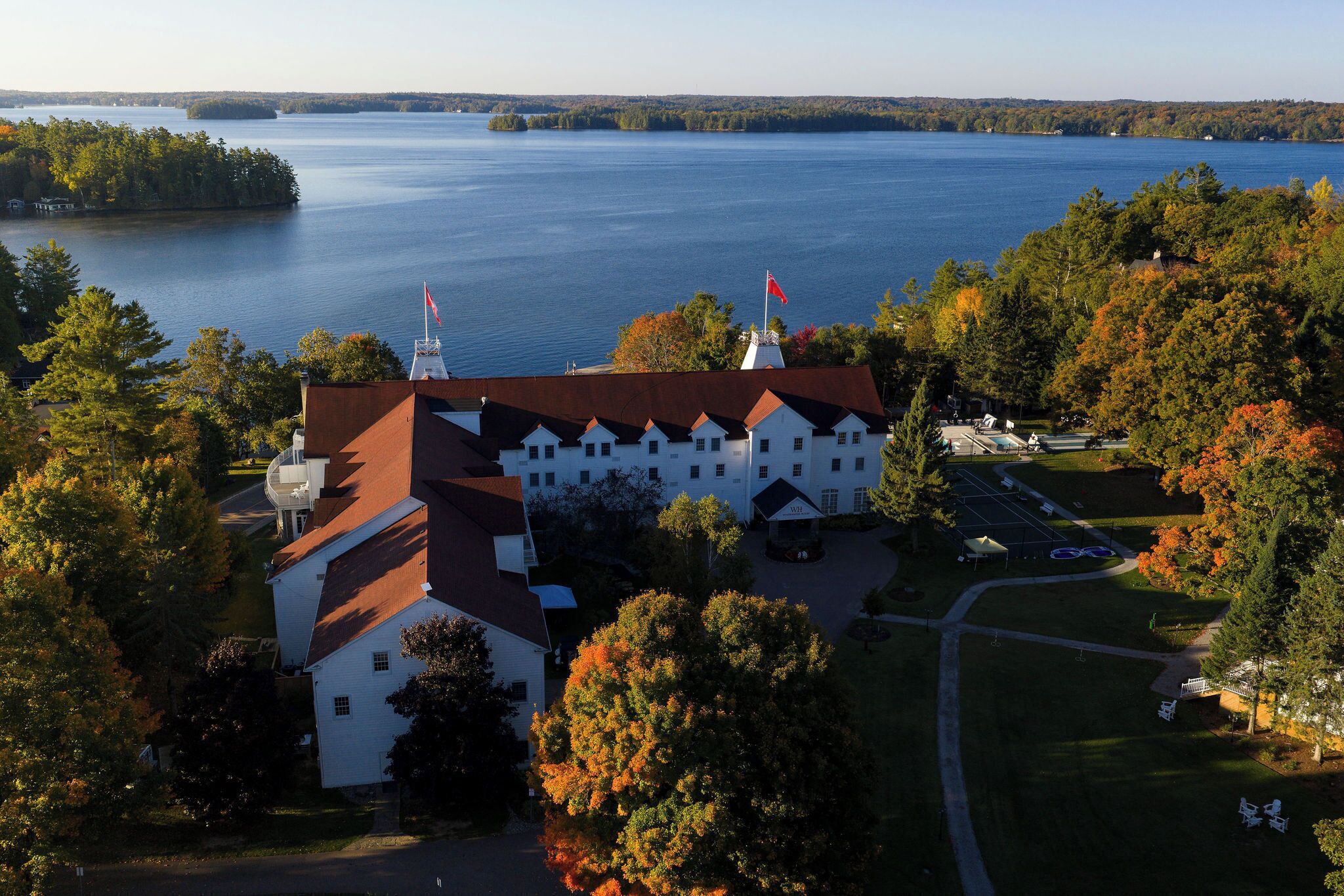 A large white building with a red roof, surrounded by trees and a lake.