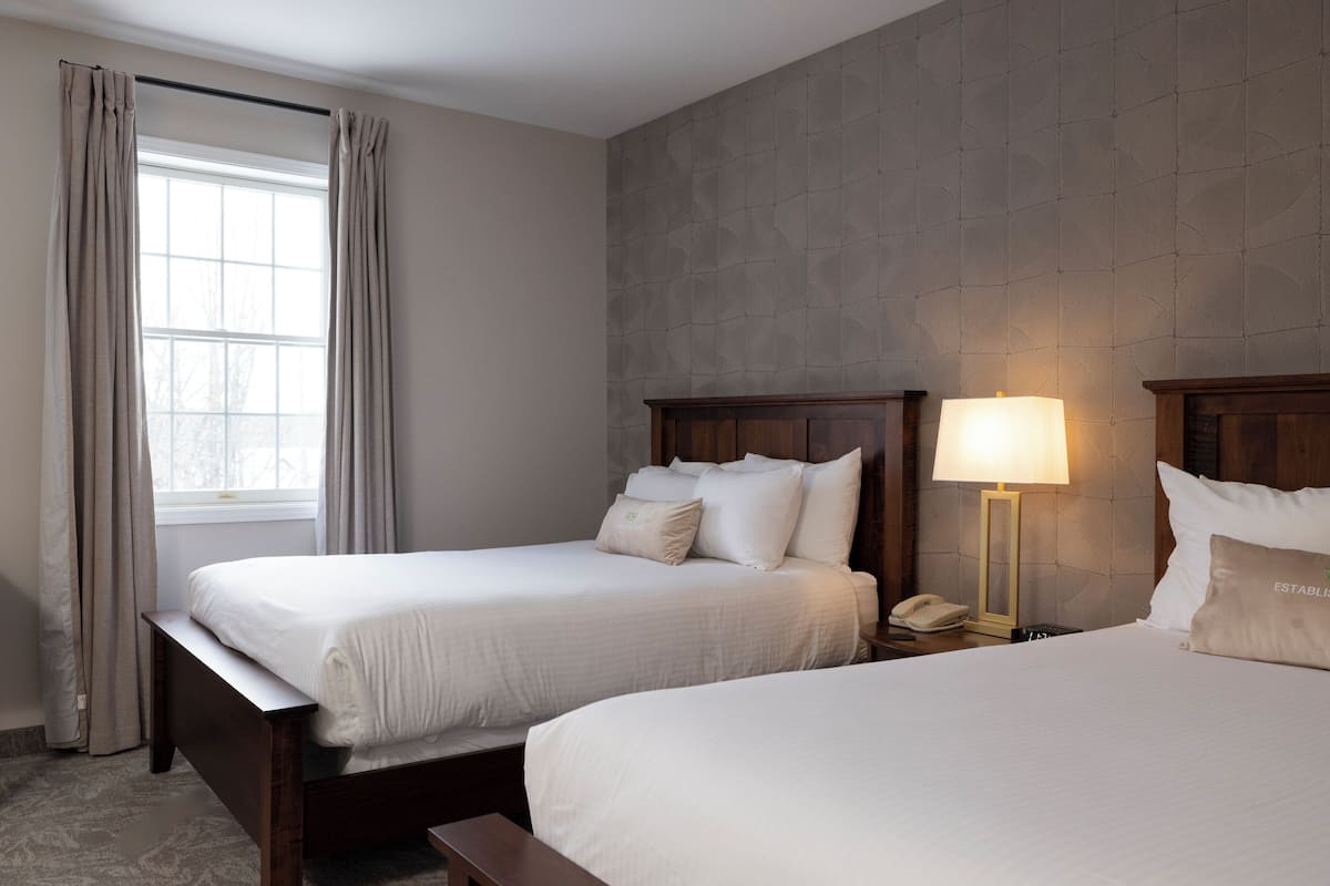 Une chambre d’hôtel avec deux lits, une fenêtre avec des rideaux, une table de chevet avec une lampe et un téléphone.