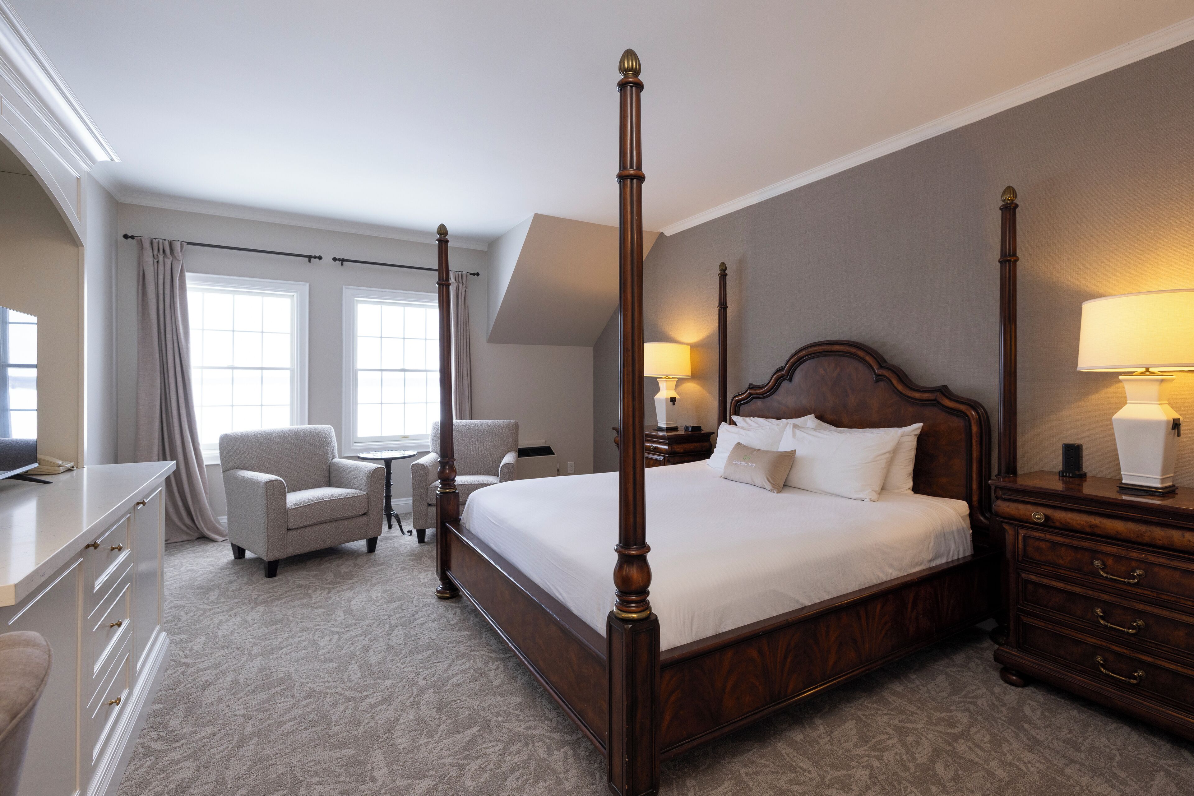 A four-poster bed with a wooden frame, a bedside table with a lamp, a chair, and a desk with drawers.