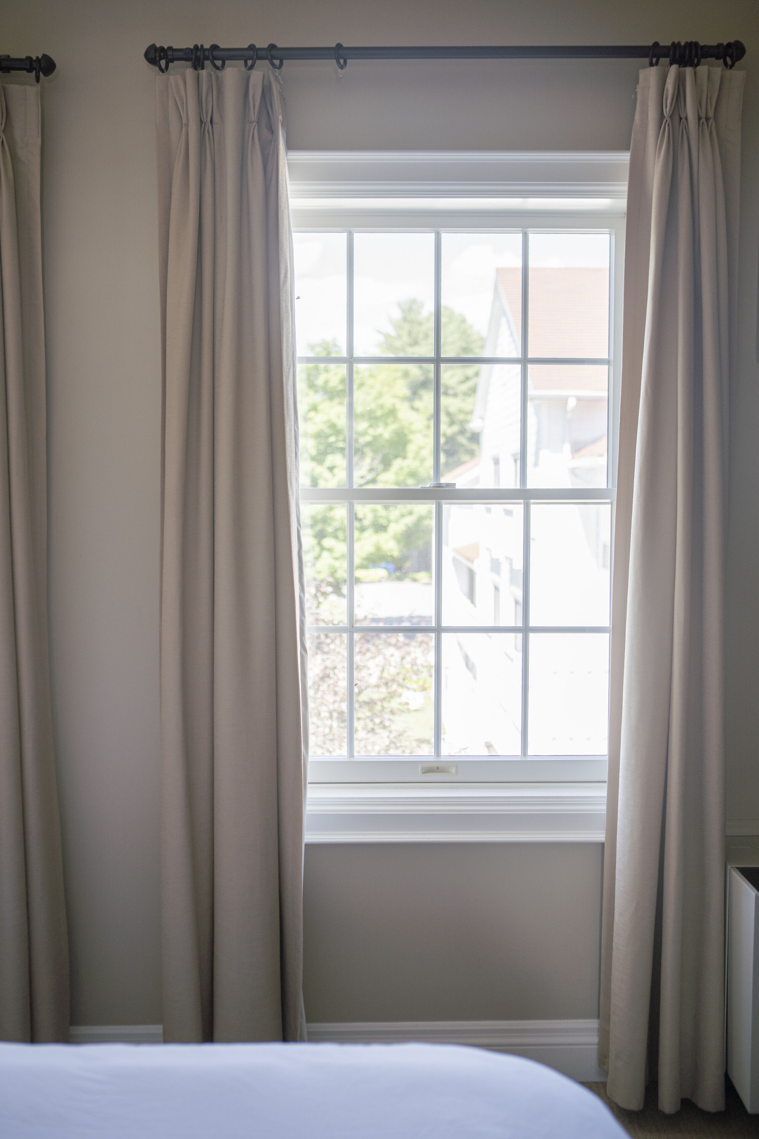 Quarto com uma grande janela, cortinas bege e cama branca.