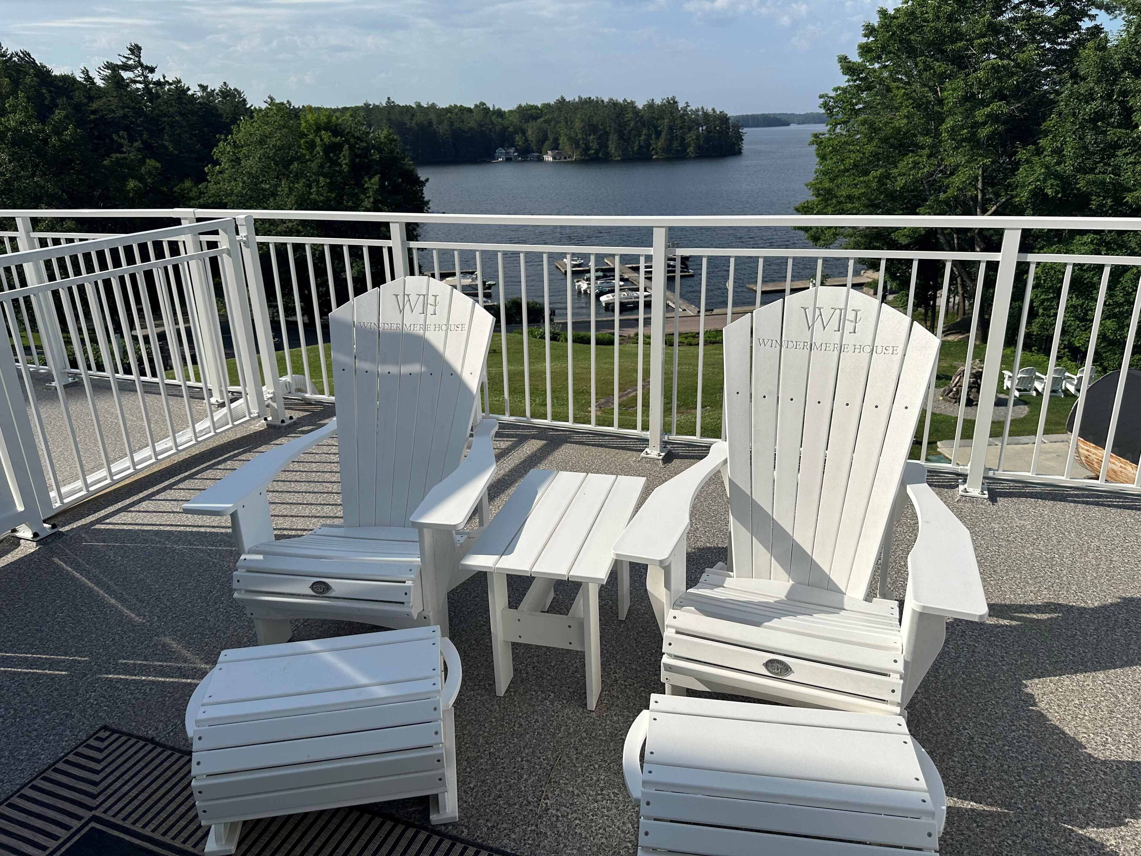 A deck with white Adirondack chairs and a table, overlooking a lake with boats and a dock.
