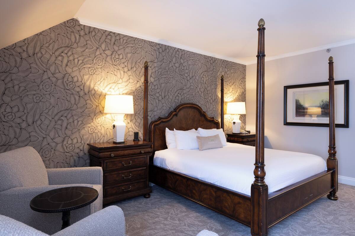 Un lit à baldaquin avec une tête de lit en bois, deux tables de chevet avec des lampes, un fauteuil à accoudoirs recouvert d’un tissu gris et un papier peint à motifs.