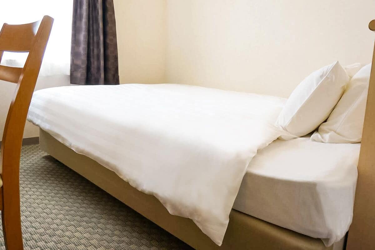 Una cama individual con colchón y almohadas blancos, una silla de madera y suelo alfombrado en una habitación con pared lisa y una ventana con cortinas.
