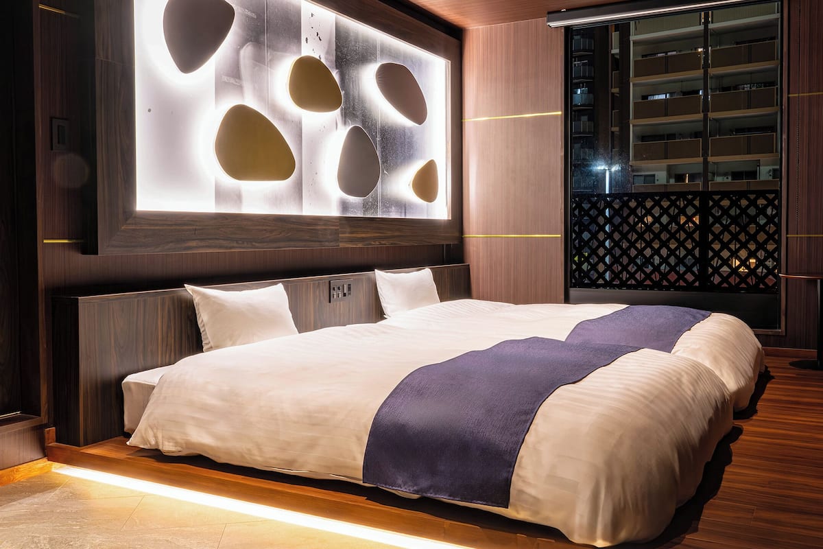 Una habitación de hotel moderna con una cama grande, un televisor montado en la pared y vistas de la ciudad a través de un ventanal amplio.