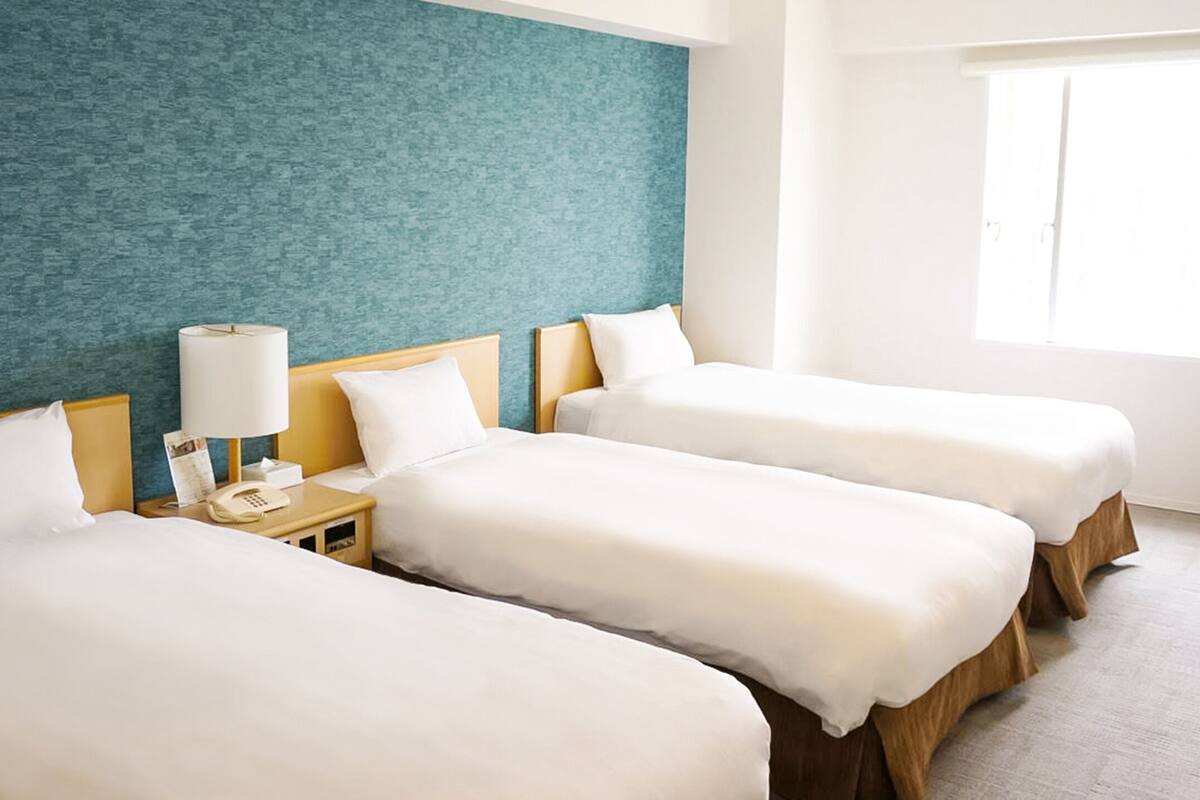 Una habitación de hotel con tres camas individuales, una mesita de noche con lámpara y una ventana.