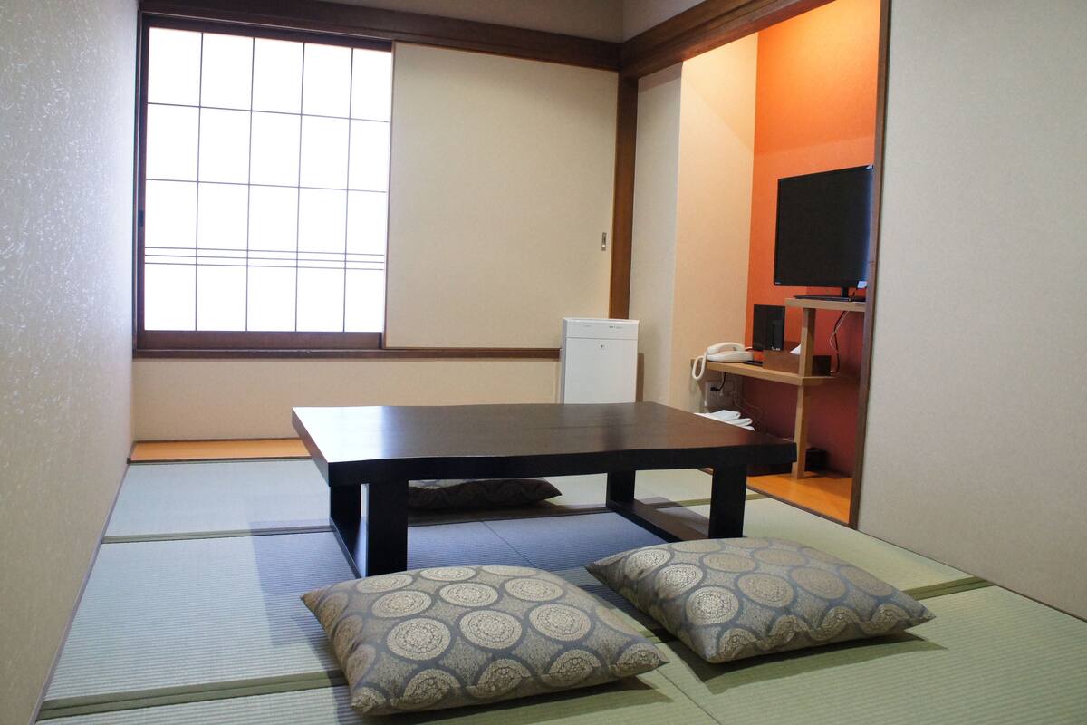 Una habitación con esteras de tatami, una mesa baja, dos cojines, un televisor y un altavoz.