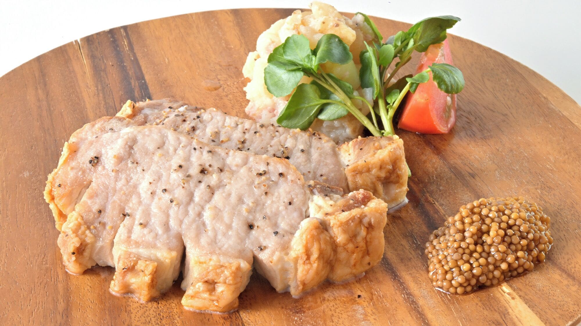 Una tabla de madera con un trozo de carne, una porción pequeña de puré de papas, una rodaja de tomate y un manojo de vegetales verdes.