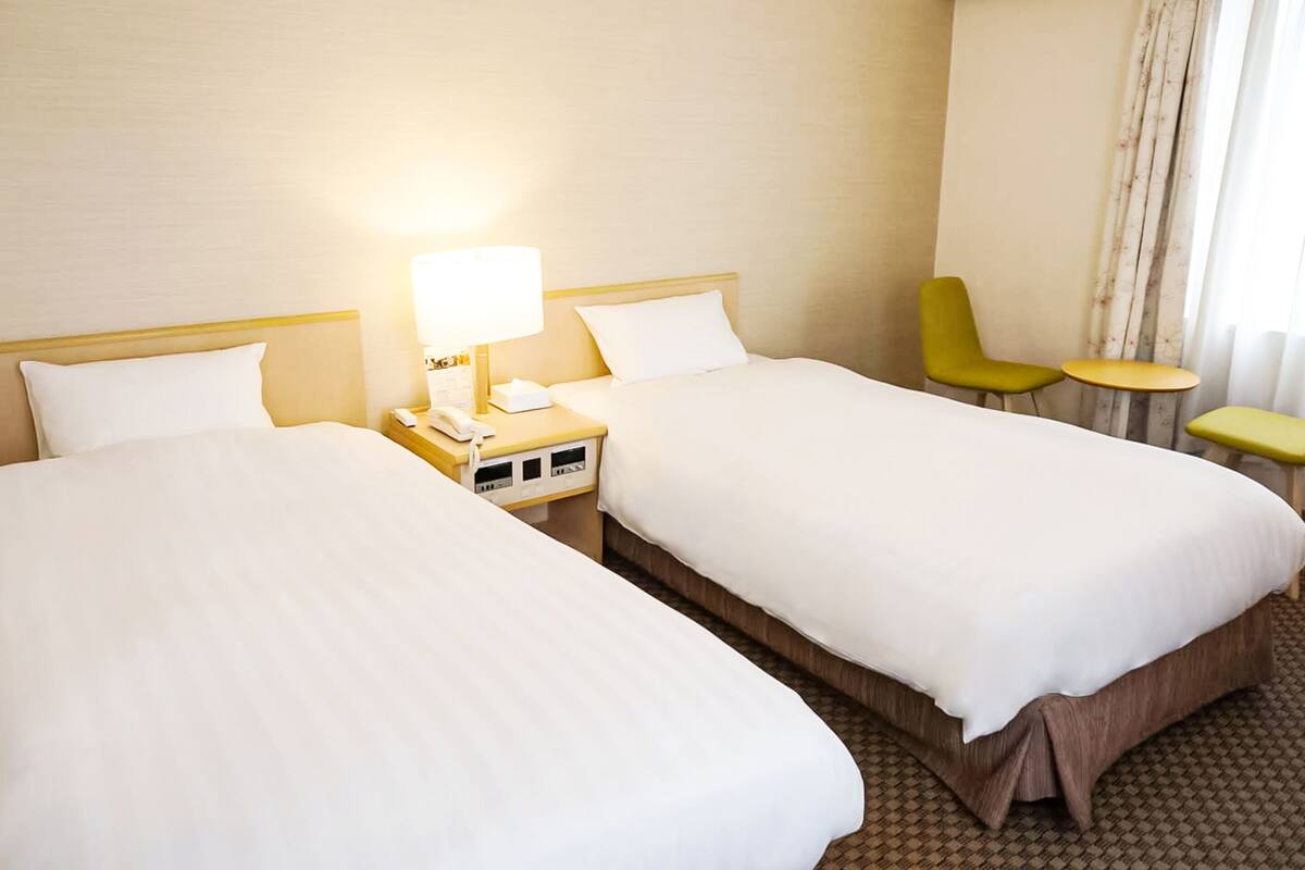 Una habitación de hotel con dos camas, una mesita de noche con lámpara, un teléfono y una pequeña mesa con sillas.
