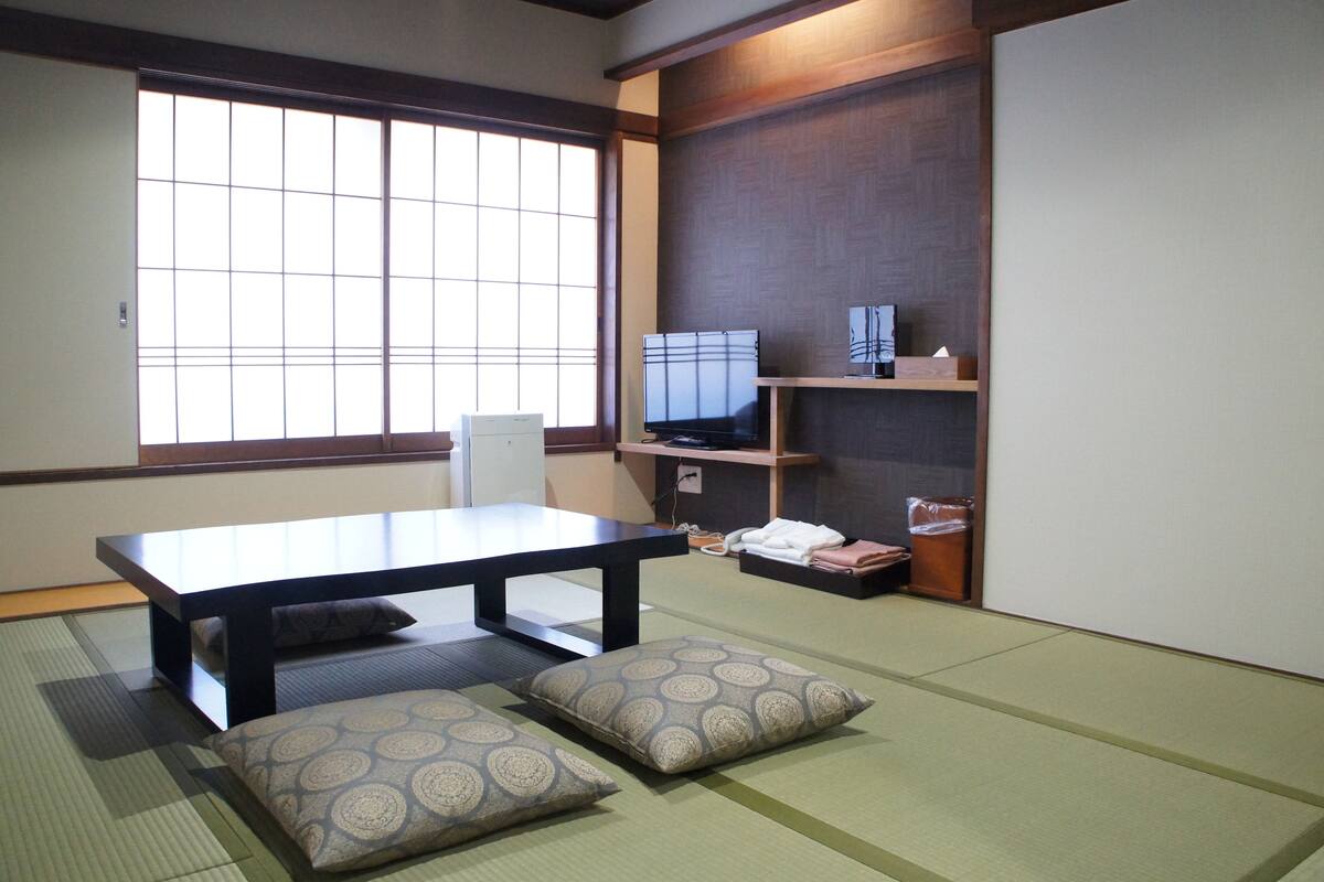 Una habitación japonesa tradicional con suelo de tatami, una mesa baja, cojines, un televisor y estanterías con objetos personales.