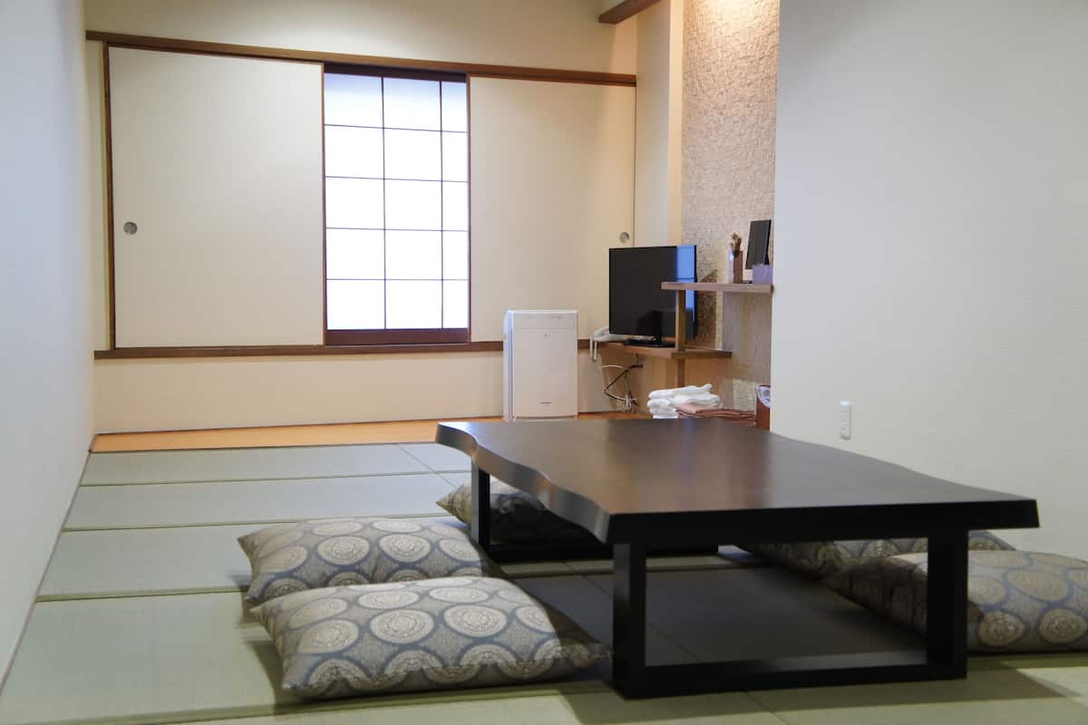 Una habitación japonesa tradicional con suelo de tatami, una mesa baja, cojines, un televisor y un refrigerador pequeño.