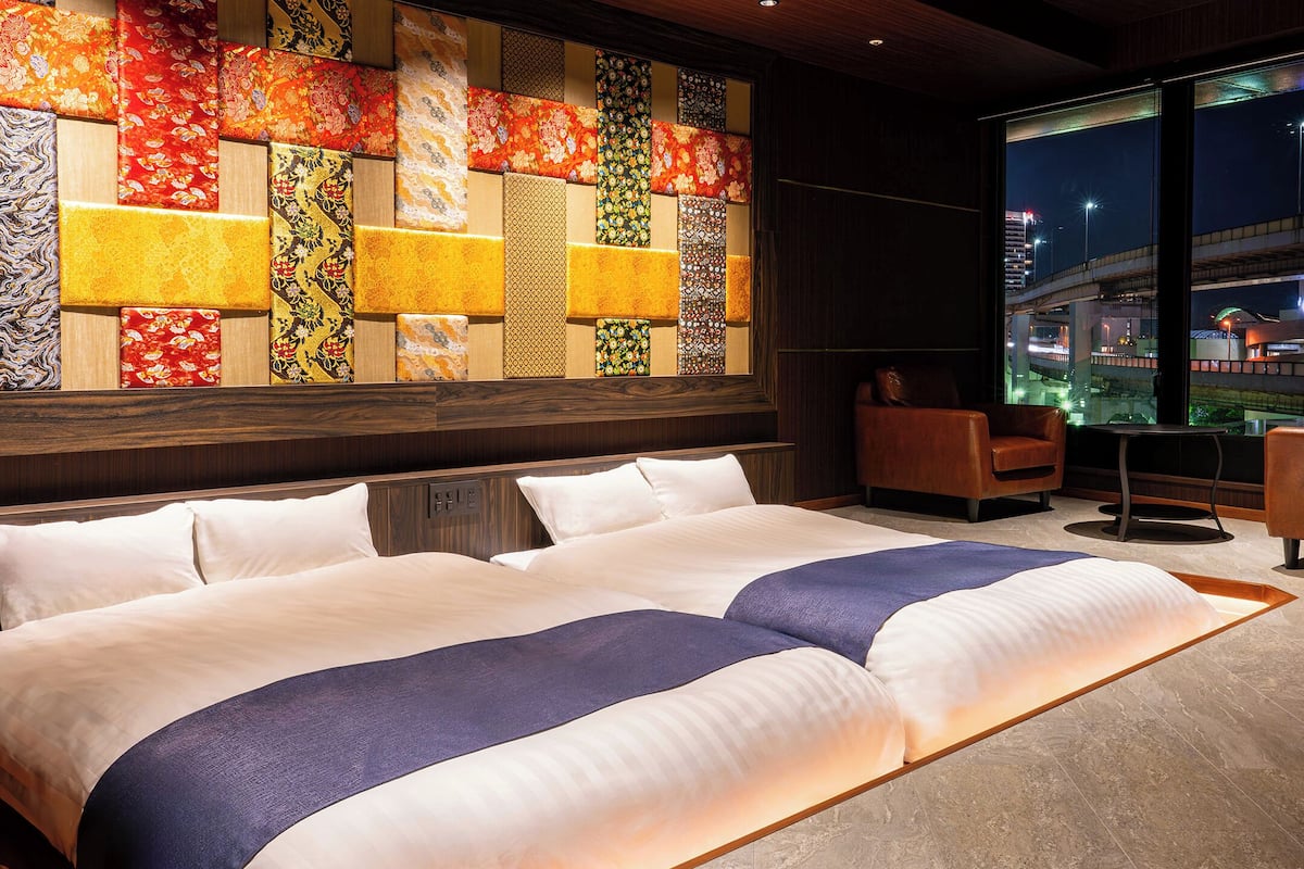 Habitación de hotel con una cama grande, dos sillones, una pared decorativa y vistas a la ciudad de noche.
