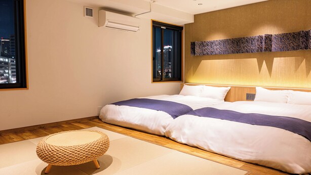 A neatly arranged hotel room with a large bed, a window with a city view, a wall-mounted air conditioner, and a woven floor mat. - Kobe PortTower Hotel (Kobe)