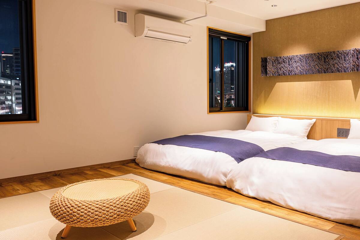 Habitación de hotel ordenada con una cama grande, un ventanal con vista a la ciudad, aire acondicionado en la pared y un tapete tejido.