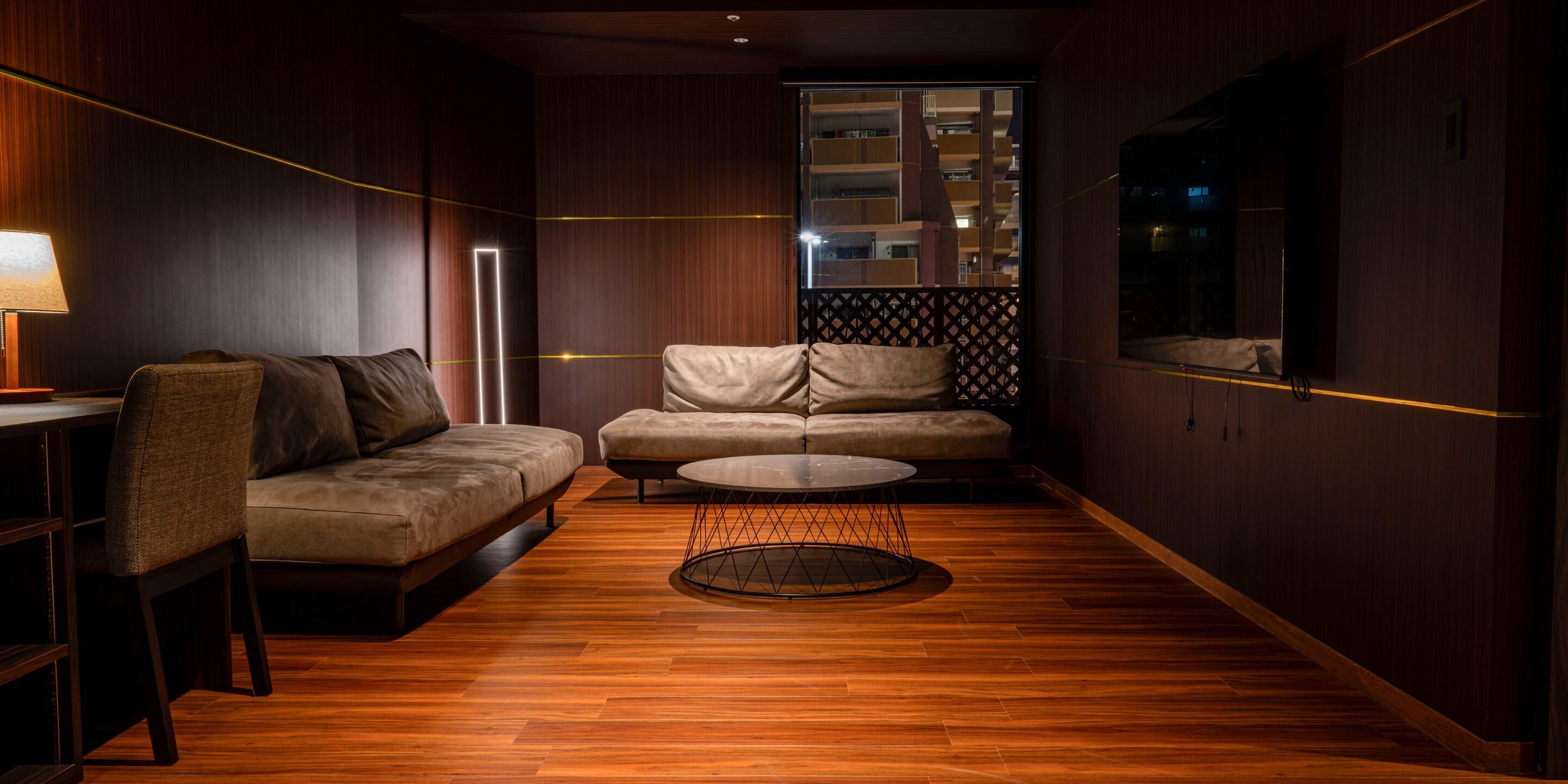 A modern living room with a sofa, armchair, and coffee table, wooden flooring, and a view of a building through a window.