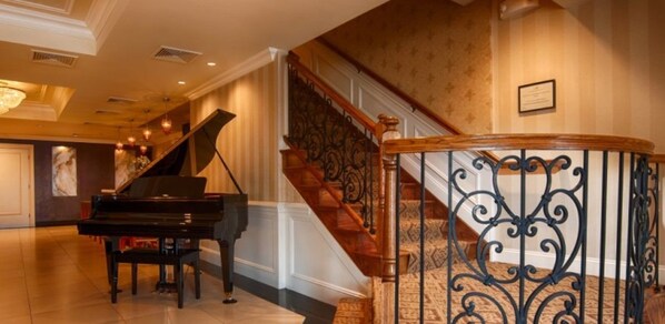 A grand piano in a spacious lobby with a wooden staircase and ornate metal railings. - American Hotel (Freehold)