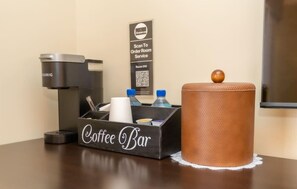 Preparación de una cafetería con una máquina de café, botellas de agua y un recipiente. Se ve un letrero de servicio a la habitación al fondo.