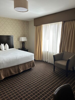 A hotel room with a large bed, a chair, a nightstand with a lamp, and a window with curtains. - American Hotel (Freehold)