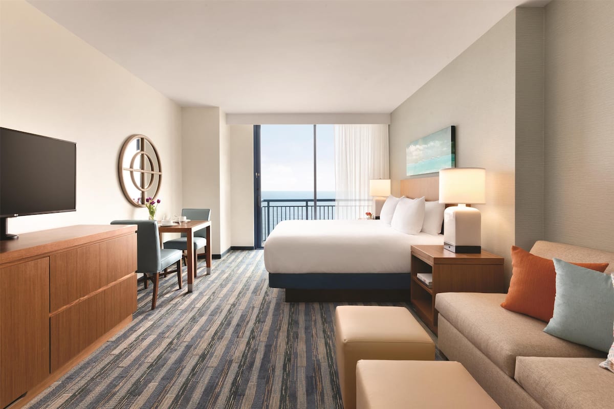 Habitación de hotel con cama, televisor, escritorio, silla, sofá y vista al cielo.