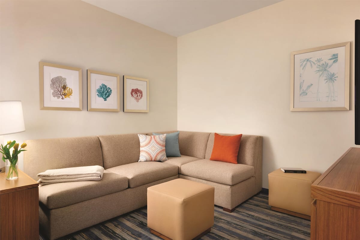 Una habitación de hotel moderna con un sofá seccional color beige, un pequeño reposapiés, una mesita de noche con una lámpara y flores, y tres obras de arte enmarcadas en la pared.