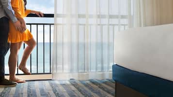 A hotel room with a bed, a view of the sea through a balcony, and a window with sheer curtains.