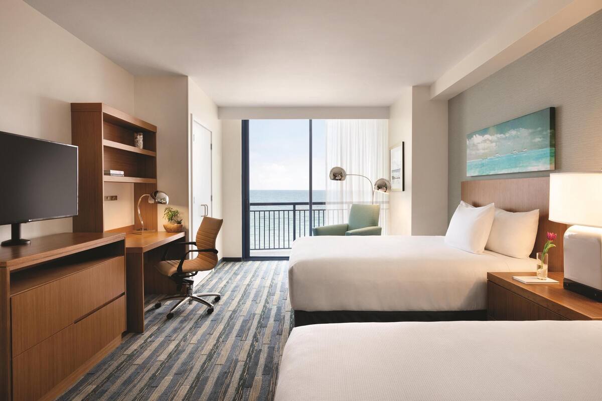 Habitación de hotel con una cama grande, un escritorio con computadora, un televisor, una silla, una mesita con lámpara y vista al océano a través de un ventanal amplio.