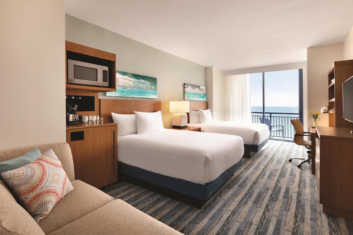 Habitación de hotel con dos camas, un sofá, un escritorio, un televisor, microondas y un amplio ventanal con vista al océano.