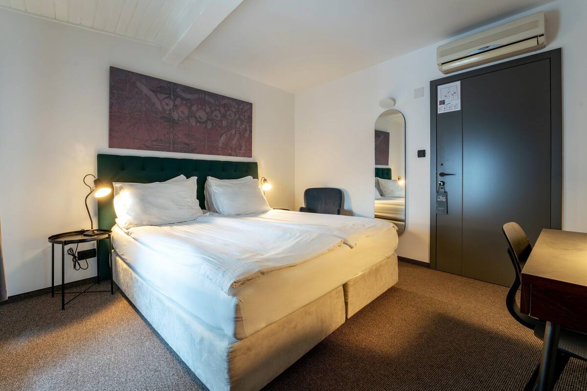 Hotellihuone, jossa on suuri sänky, yöpöytä lampun kanssa, tuoli, työpöytä ja ilmastointilaite.