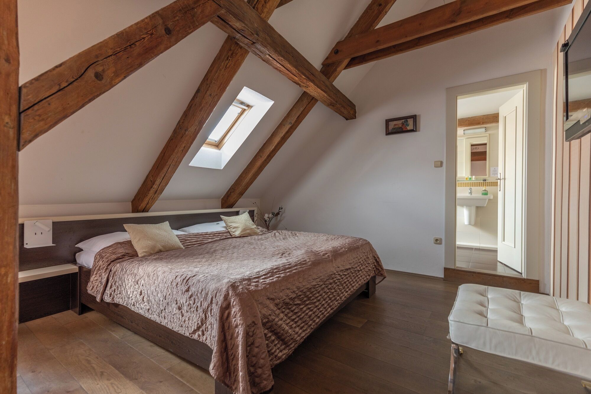 A bedroom with a sloped ceiling, a bed with a textured blanket, a small window, and a view of a bathroom.