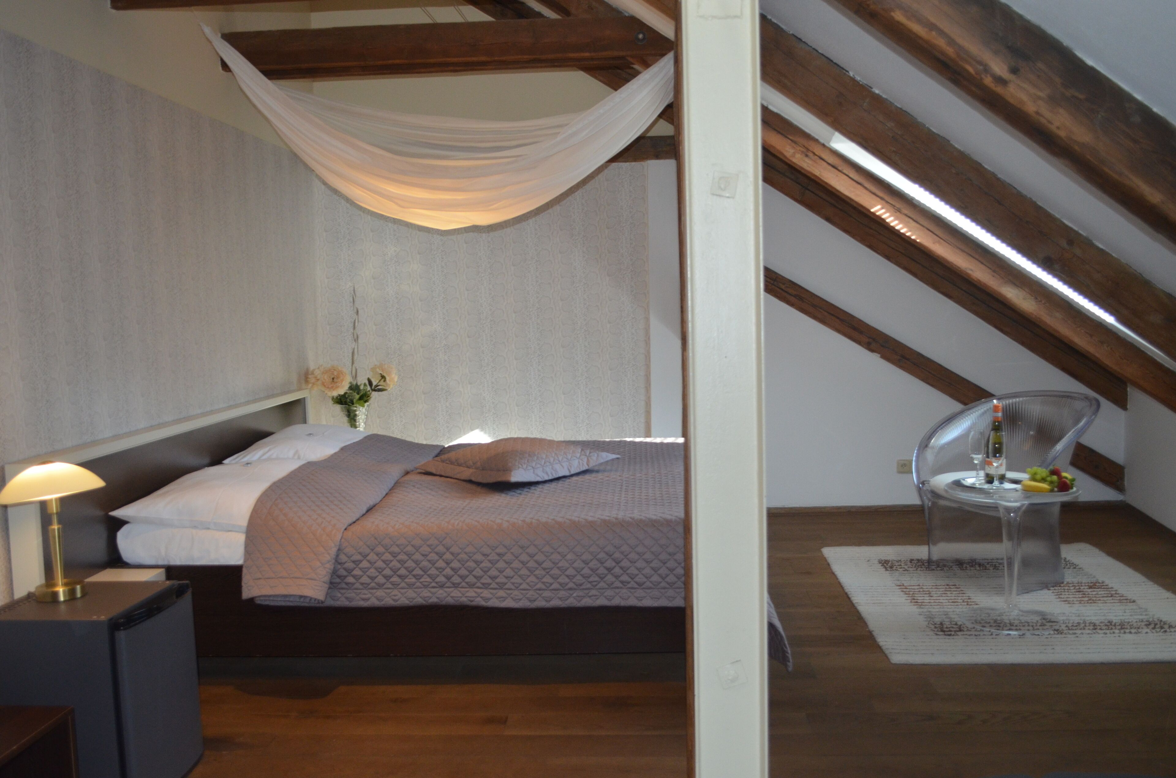 Een slaapkamer met een bed, nachtkastje, lamp, stoel en een klein tafeltje met fruit en een fles.