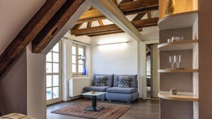 A cozy loft bedroom with exposed wooden beams, a gray sofa, a small table, and a built-in shelf with glasses. - Hotel Peregrin (Cesky Krumlov)