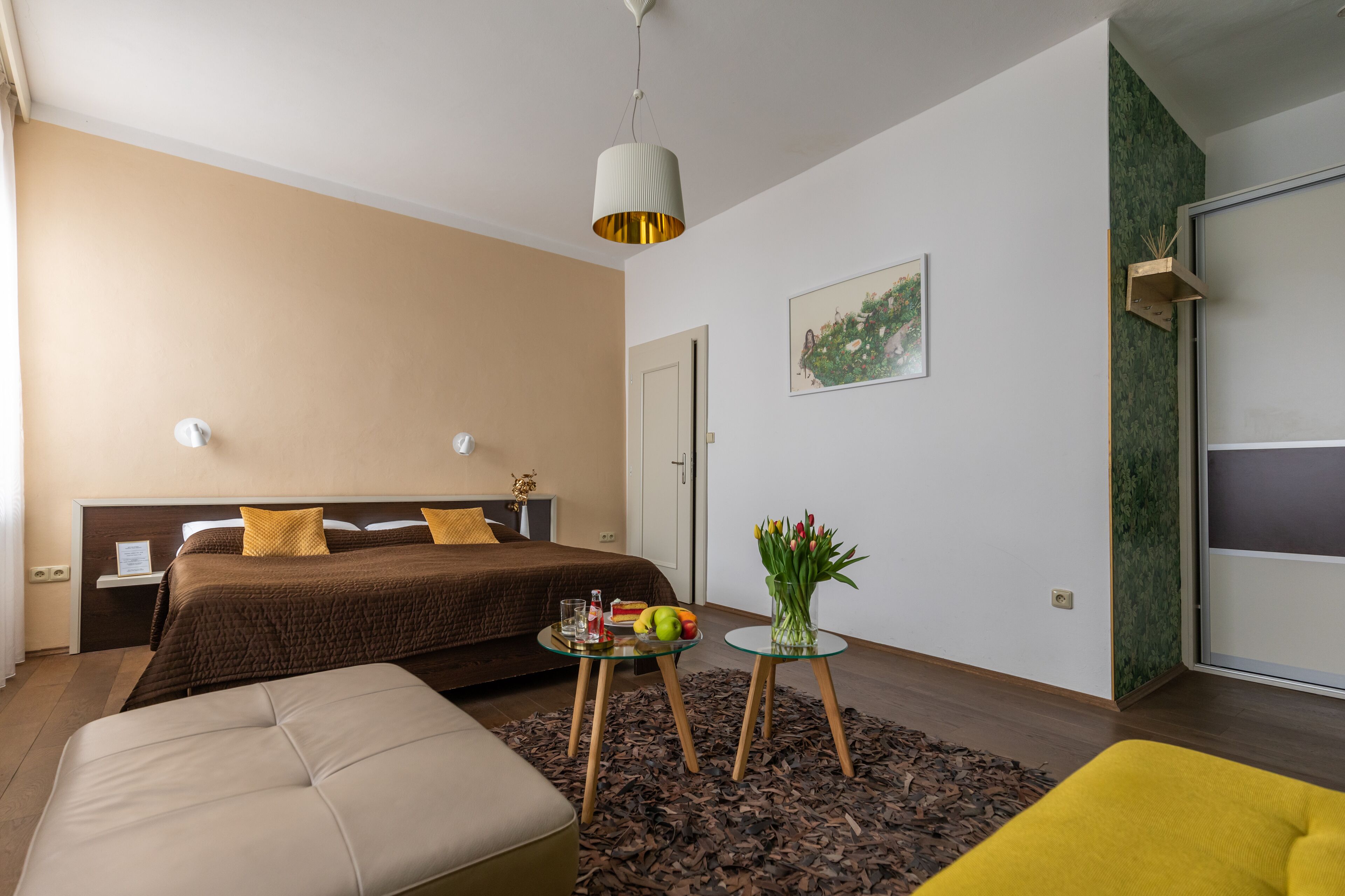 A modern bedroom with a large bed, bedside tables, a small table with fruit and flowers, and a yellow armchair.