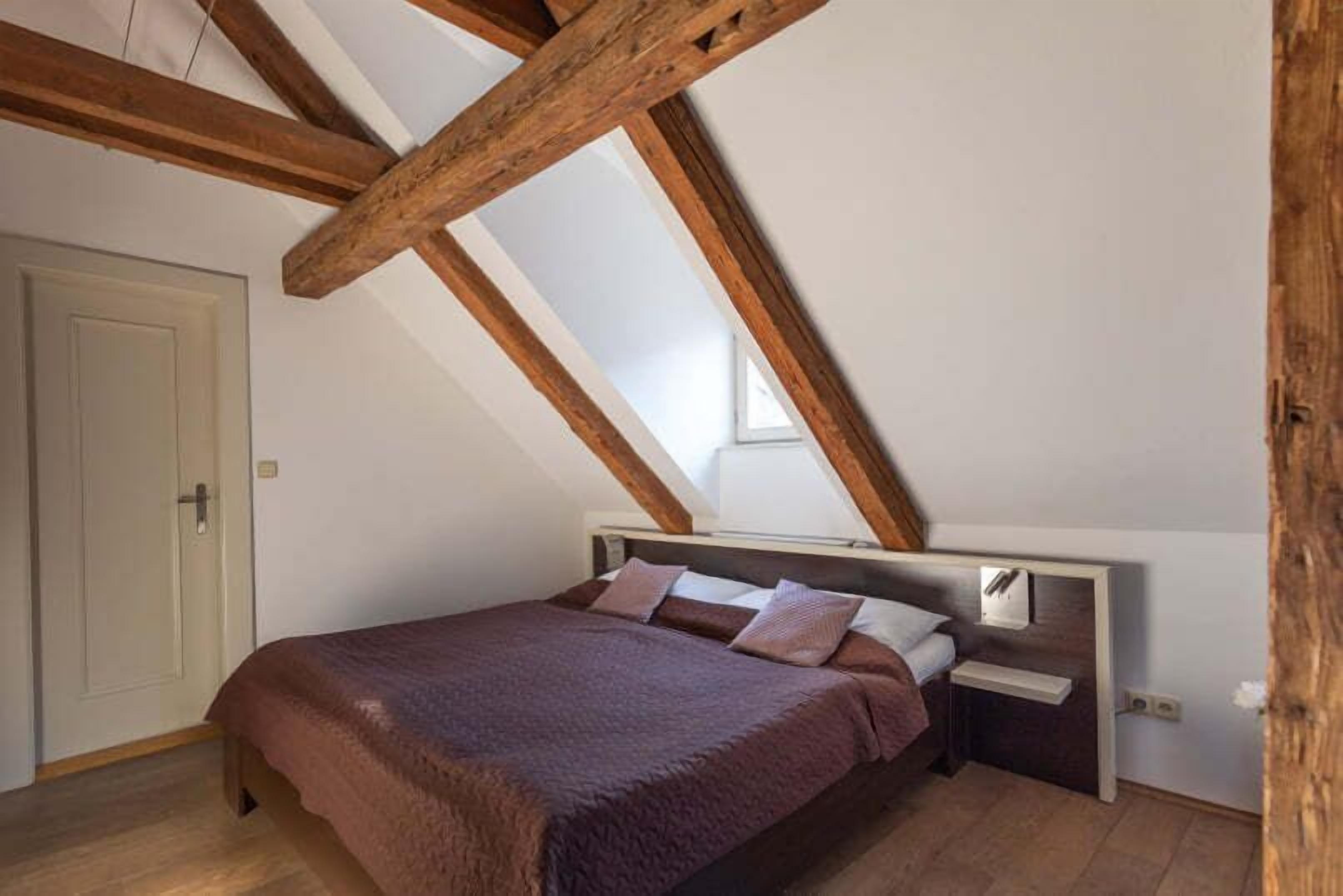 A bedroom with a sloped ceiling, wooden beams, a bed with a headboard, a bedside table, and a door.