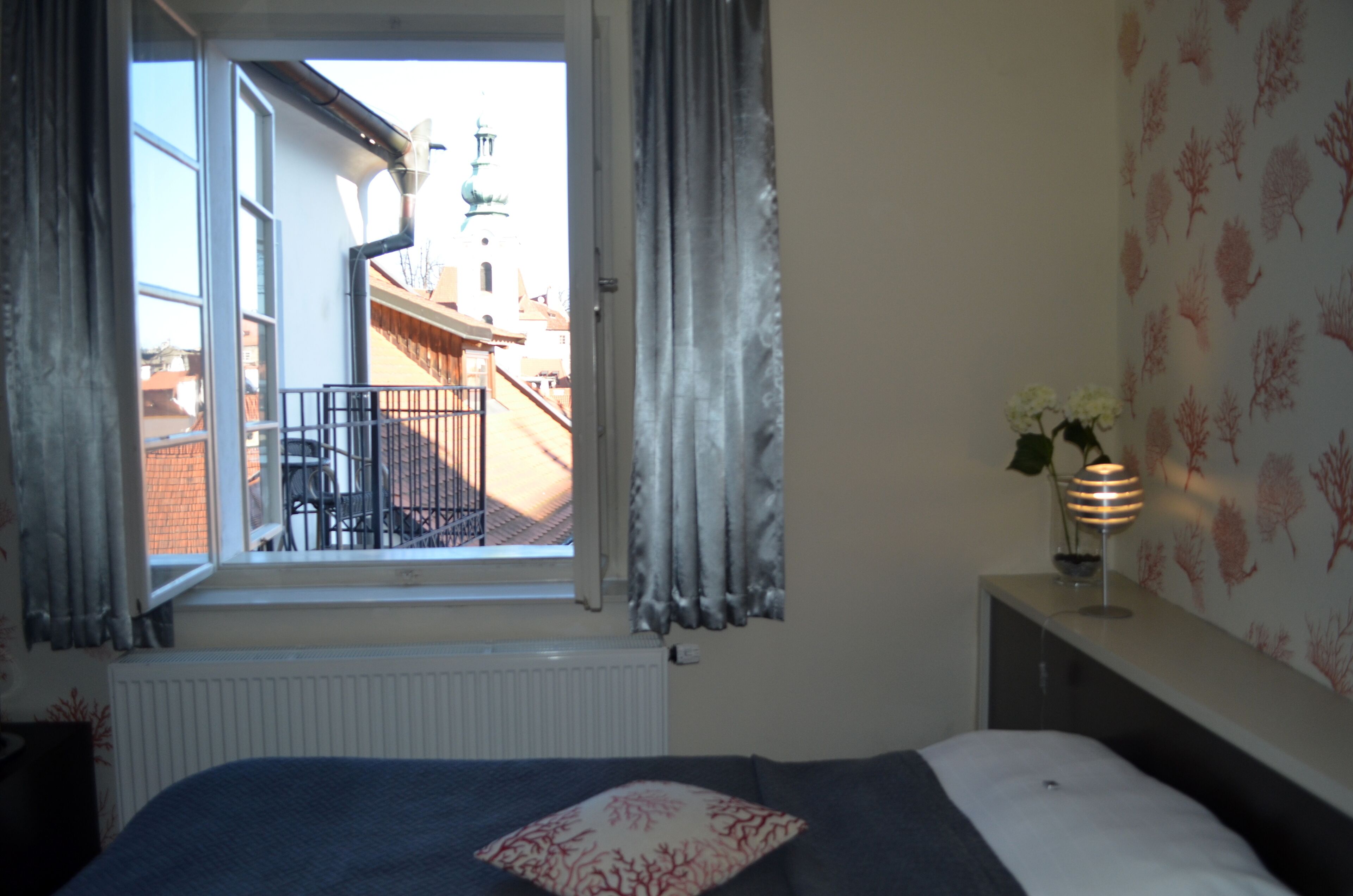 A bedroom with a bed, a window with curtains, a radiator, a nightstand with a lamp, and a wall with a leaf pattern.