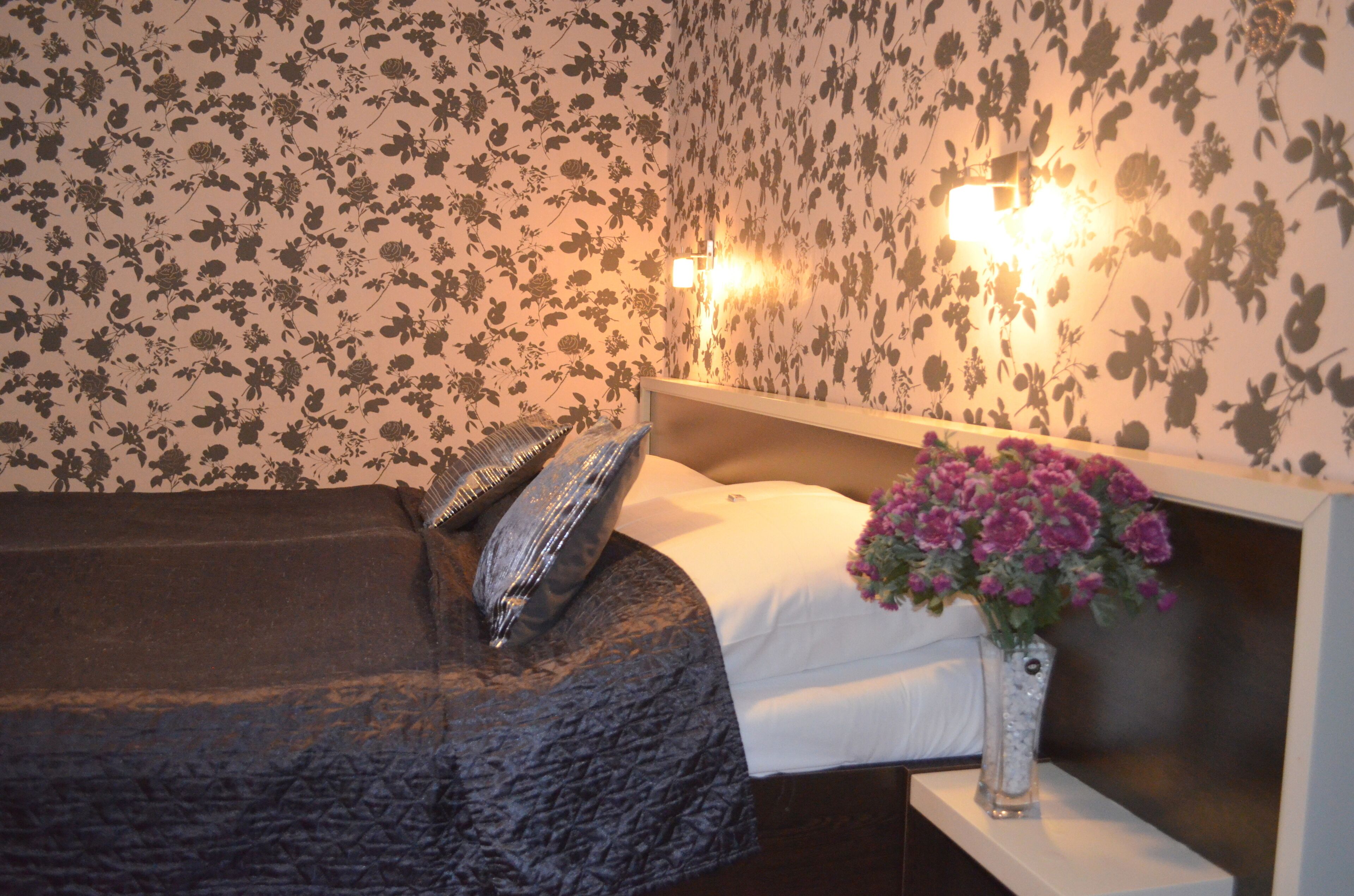 A bedroom with a bed, pillows, a vase with flowers, and floral wallpaper.
