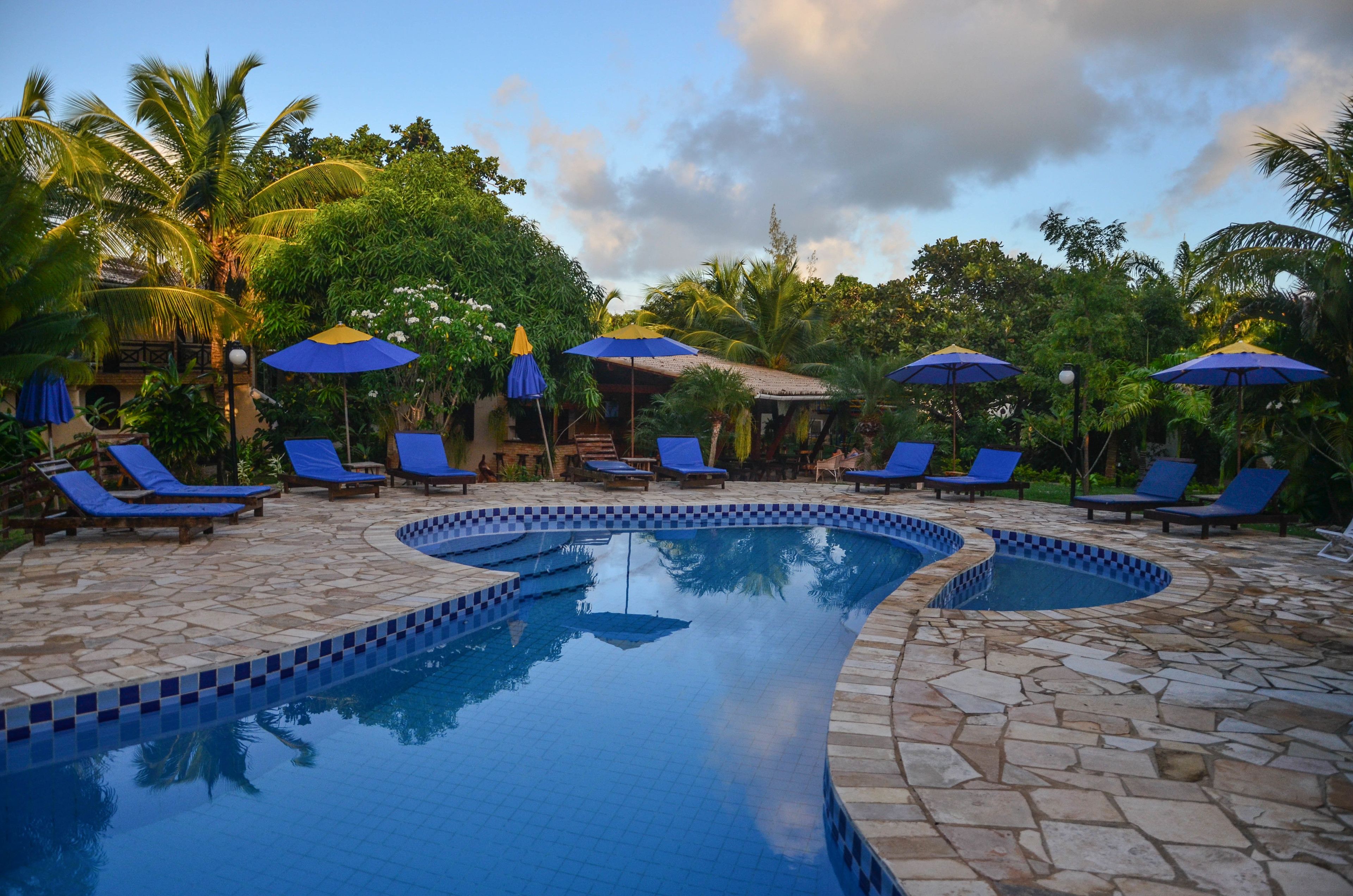 Un área de piscina con sillas reclinables azules y sombrillas, rodeada de palmeras y un deck empedrado.
