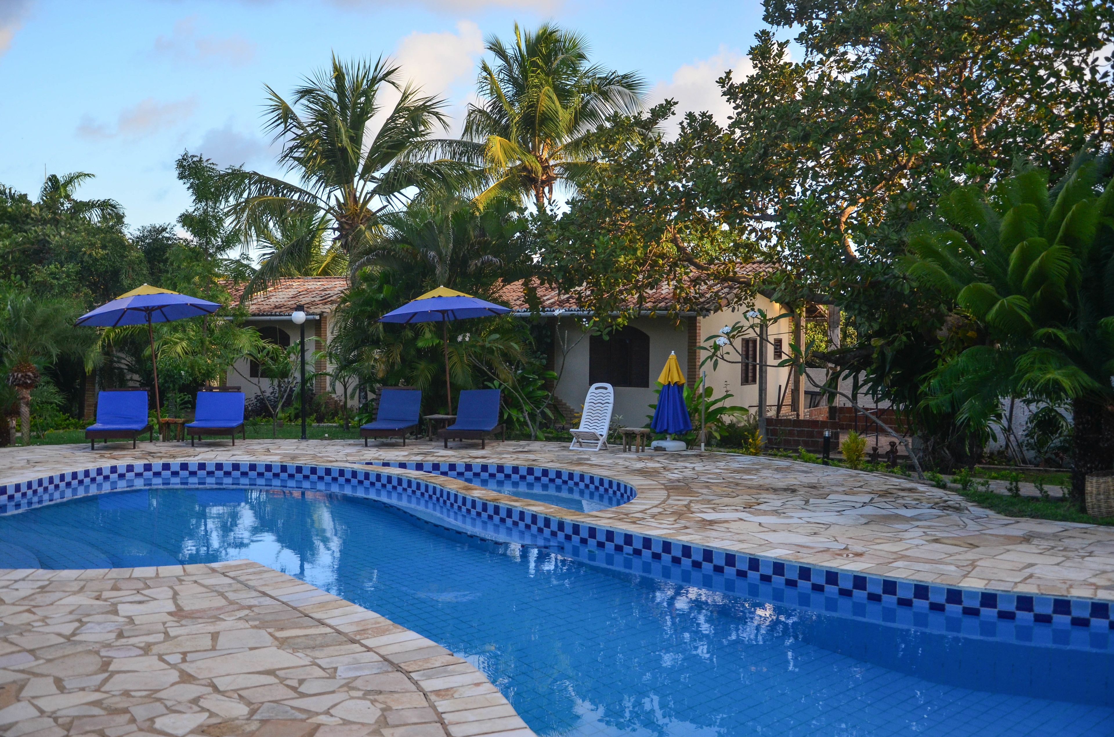 Una piscina con baldosas azules formando un diseño de cuadros, rodeada de sillas de descanso y sombrillas, con palmeras y un edificio al fondo.