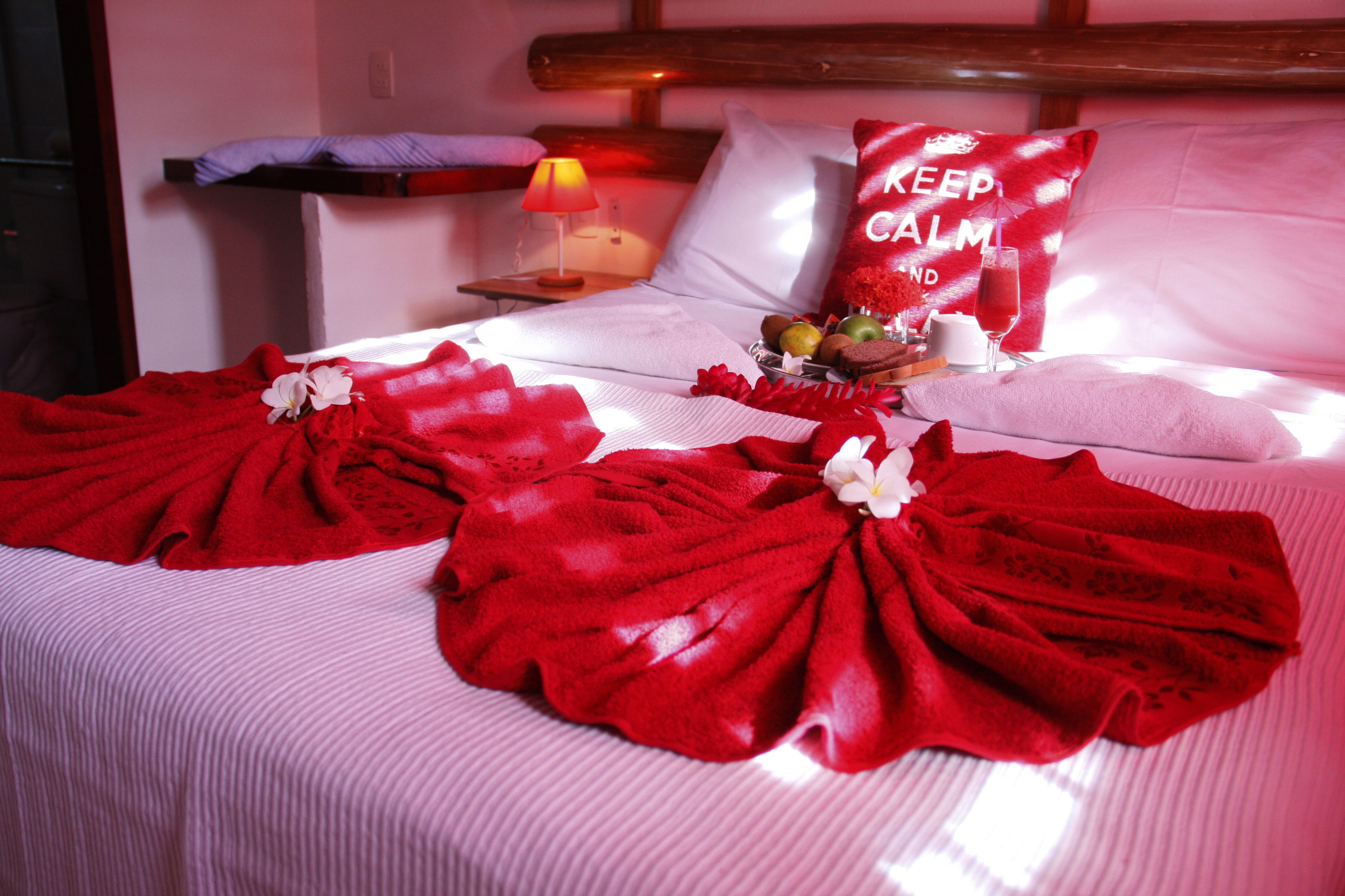 Una habitación de hotel con una cama bien hecha, un almohadón con la inscripción "KEEP CALM AND", una mesita de noche con una lámpara y una bandeja de frutas y bombones.