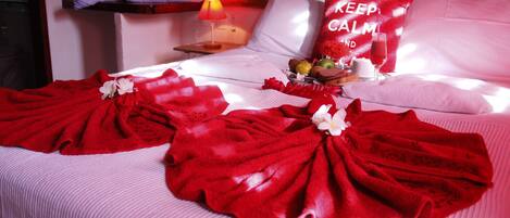 Una habitación de hotel con una cama bien hecha, un almohadón con la inscripción "KEEP CALM AND", una mesita de noche con una lámpara y una bandeja de frutas y bombones.