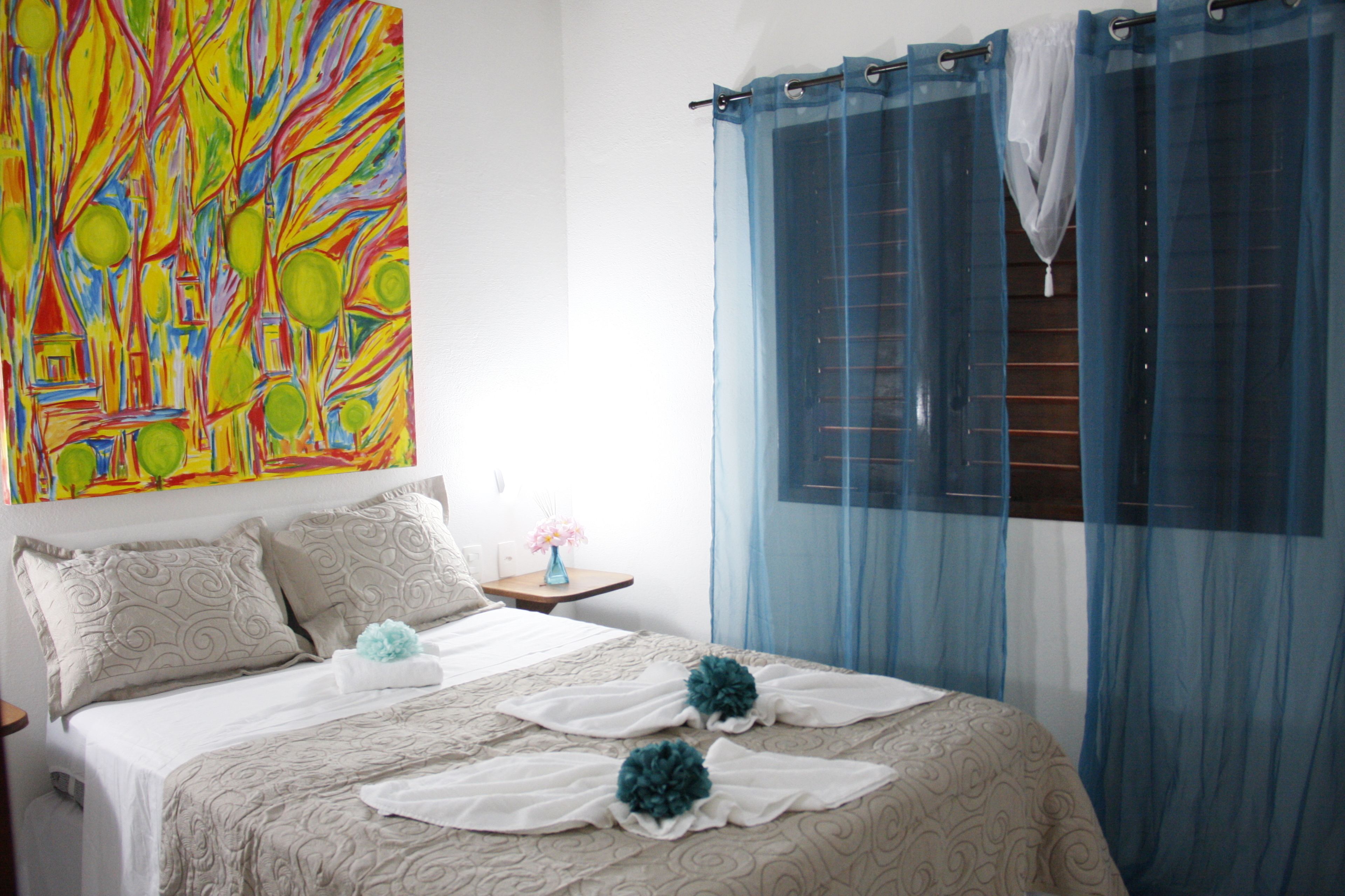 Un dormitorio con cama, almohadas, una mesita de noche, un jarrón con flores y un cuadro abstracto de colores en la pared.