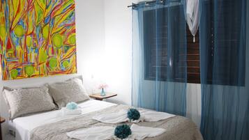 Un dormitorio con cama, almohadas, una mesita de noche, un jarrón con flores y un cuadro abstracto de colores en la pared.